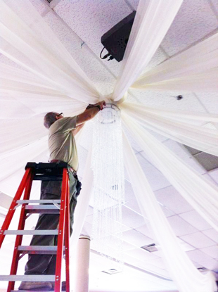 Ernesto up in the ladder installing white chiffon ceiling treatments and a decorative crystal chandelier for a wedding in Orlando, FL. Photo by W Drapings.