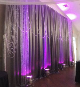 Custom backdrop in silver satin with decorative strings of crystal beads and purple LED up-lights. Photo, drapes and accessories by W Drapings.